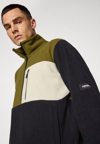 Fleece pulóver v olivovo zelenej, krémovej a námorníckej. Obsahuje polovičný zips na krku, bočné vrecká a nášivku loga na rukáve. Textúrovaná látka.