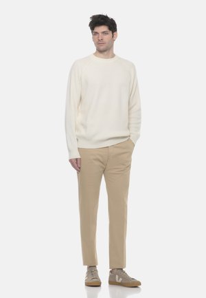 Uomo con maglione a maglia color crema, pantaloni beige e sneakers beige, in piedi con una mano in tasca contro uno sfondo bianco semplice.