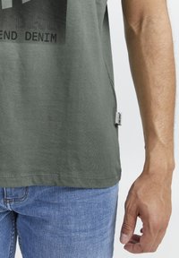 Olivegrønn bomulls T-skjorte med svart trykk på forsiden, med et merke på sidesømmen. Matchet med blå denimjeans.