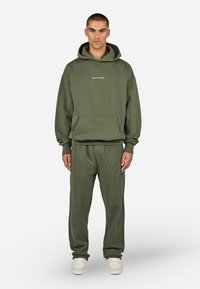 Zelená bavlnená mikina s kapucňou a prednou kapsou, s vyšitým logom. V kombinácii s nohavicami so vzorovaným rebrovaním a bielymi teniskami.