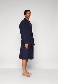 Navy blue, textured bathrobe with a shawl collar and belt, knee-length, featuring a woven pattern and long sleeves. Bare feet visible.