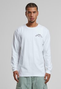 Weißes Langarm-T-Shirt aus Baumwolle mit einem kleinen schwarzen Logo auf der Brust. Glatte Textur, klassischer Rundhalsausschnitt und taillierte Ärmel.