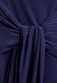 Mørkeblåt stof samlet og bundet i en knude, der skaber bløde folder og plisseringer på glat materiale.