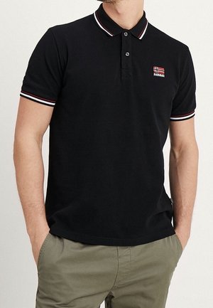 Mann trägt schwarzes kurzärmeliges Poloshirt mit rotem, weißem und schwarzem Besatz am Kragen und an den Ärmeln, kombiniert zu olivgrünen Hosen.