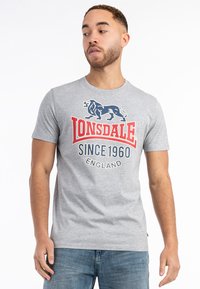 Grå kortærmet t-shirt med et blåt løvegrafik og rød tekst "Lonsdale Since 1960 England" på fronten. Casual fit.