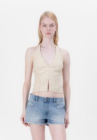 Top corto con cuello halter en un estampado de cuadros en tonos beige y blanco, que presenta un escote en V profundo y una abertura en la parte delantera, combinado con unos pantalones cortos de mezclilla azul.