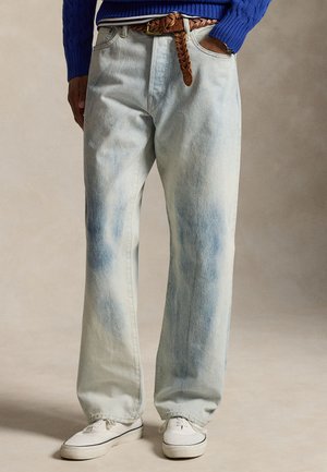 Persoană purtând blugi albastru deschis decolorați cu brâu din piele maro împletită, pulover albastru, adidași albi, mâinile în buzunare, fundal neutru.