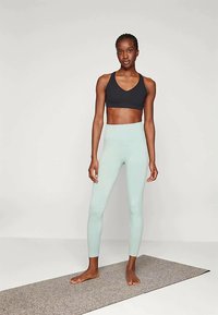 Melnā sporta krūšturis un gaiši teal krāsas legingi, izgatavoti no elastīga auduma, pieguļošs dizains, gluda tekstūra un minimālisma stils. Stāv uz pelēka paklāja.