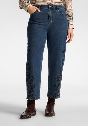 Persona che indossa jeans in denim blu con ricamo floreale nero sulle gambe inferiori, abbinati a calzini marroni e mocassini marrone scuro.