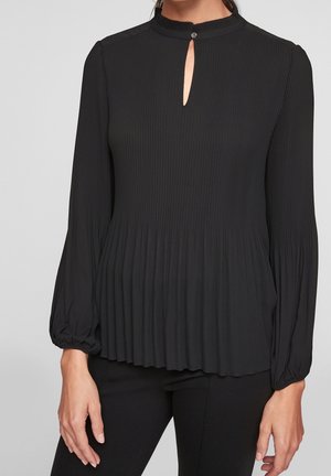 Vrouw draagt een lange mouwen, geplooide zwarte blouse met een klein sleutelgat aan de voorkant en knoopsluiting bij de halslijn.