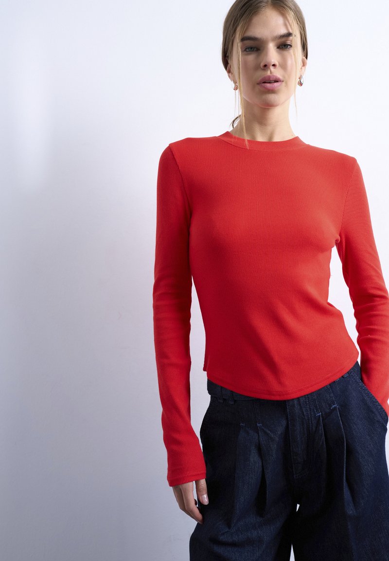 Γυναίκα που φοράει εφαρμοστή κόκκινη μπλούζα με μακριά μανίκια και χαλαρό σκούρο τζιν παντελόνι, στέκεται μπροστά σε απλό ανοιχτό φόντο.