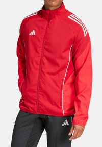 Rode sportjack met een ritssluiting aan de voorkant, witte accenten, en drie witte strepen op de schouders, gemaakt van lichtgewicht stof.