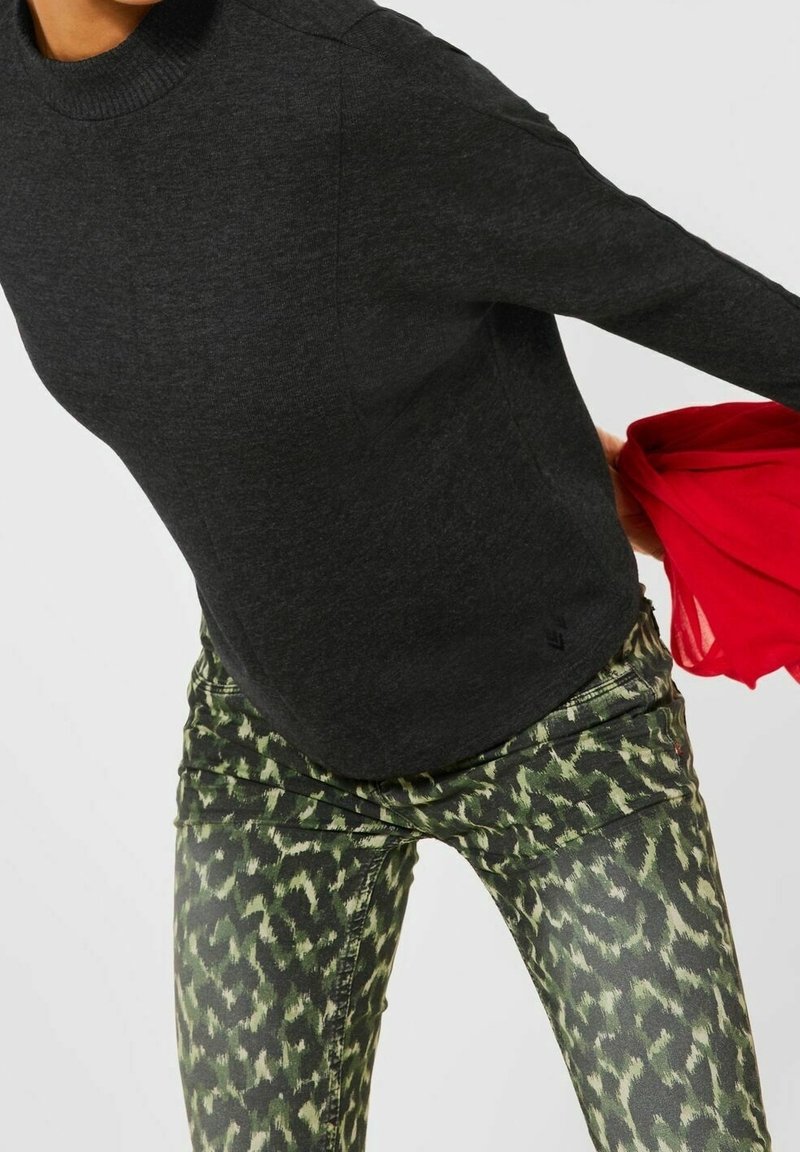 Top de manga larga gris oscuro con cuello redondo, combinado con pantalones ajustados verdes con un patrón que presenta un sutil estampado de animales. Tela roja parcialmente visible.