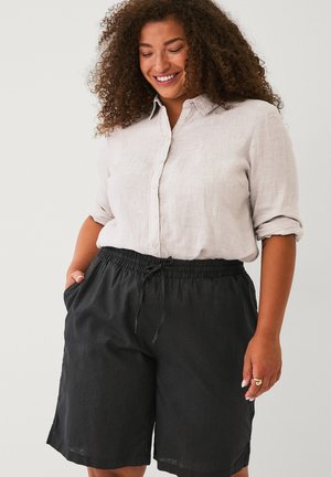 Smilende kvinde med krøllet hår iført en lys beige skjorte med knapper og ærmerne rullet op samt sorte shorts med snor i livet og hænderne i lommerne.