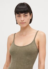 Débardeur côtelé vert olive avec de fines bretelles, présentant un col scoop et un tissu texturé. Le design sans coutures améliore la coupe ajustée.