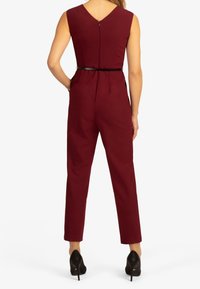 Mono burdeos con cintura ajustada, espalda en forma de V y cinturón en la cintura. Presenta piernas rectas y está combinado con zapatos de tacón negros.