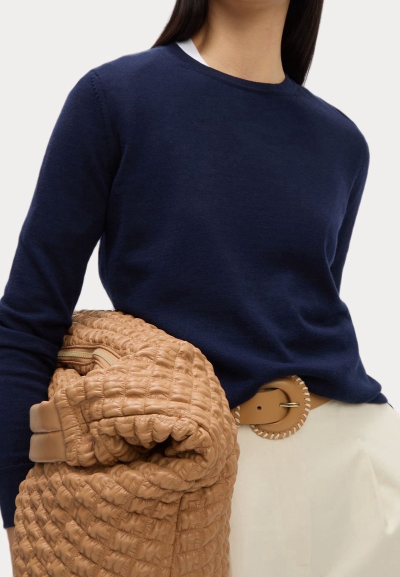 Femme portant un pull bleu marine et un pantalon crème avec une ceinture tressée beige, tenant un grand sac matelassé beige plié devant un fond blanc.