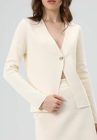 Blazer couleur crème en tissu texturé, avec un col en V, des manches longues et une fermeture à un seul bouton argenté à la taille.