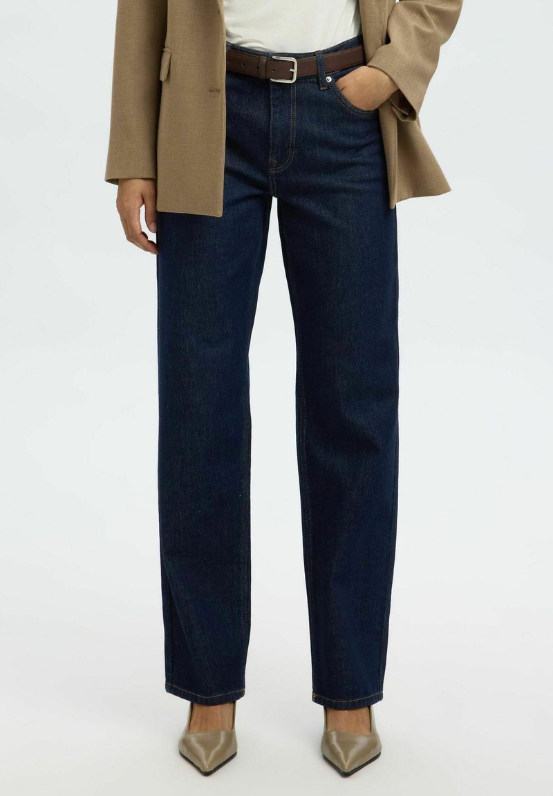 Frau trägt dunkelblaue Jeans mit geradem Bein, beigen Blazer, weißes Top, braunen Gürtel und beige spitze Absätze, steht mit einer Hand in der Tasche.