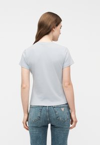 Νεαρή γυναίκα με μακριά καστανά μαλλιά γυρισμένη προς τα πίσω, φοράει ανοιχτό γκρι κοντομάνικο πουκάμισο και μπλε τζιν με πίσω τσέπες.