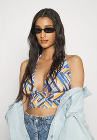 Top crop halter colorido com padrões geométricos em azul, laranja e cinza; combinado com uma jaqueta de jeans clara e calças de cintura alta.