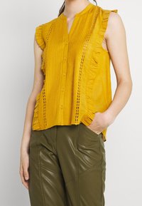 Žlutá blůza s volánovými rukávy, vertikálními sklady a předními knoflíky. Obsahuje složité krajkové detaily na boku.