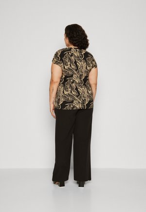 Femme aux cheveux bouclés se tenant face à un mur, portant un haut noir avec des motifs de feuilles beiges et un pantalon noir ample avec des talons blocs.