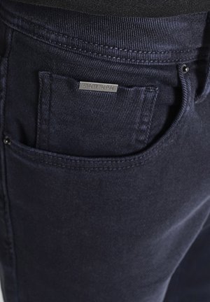 Gros plan sur la poche d'un jean bleu marine avec des coutures visibles et une petite étiquette en métal de la marque au-dessus de l'ouverture de la poche.