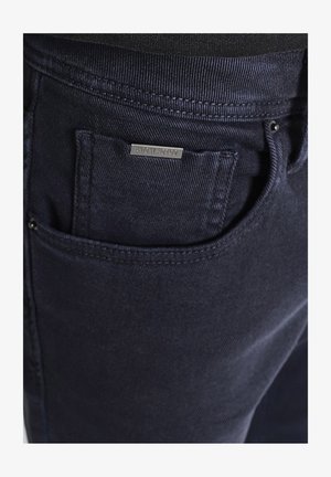 Close-up van een marineblauwe jeanszak met zichtbare stiksels en een klein metalen merketiket boven de opening van de zak.