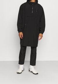 Schwarzer, leichter Anorak mit Reißverschluss vorne, Kapuze und zwei Seitentaschen; mit einem Logo-Aufnäher auf der Brust; kombiniert mit schwarzen Hosen und weißen Schuhen.