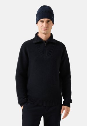 Man draagt een zwarte trui met halve rits, bijpassende zwarte broek en een donkerblauwe gebreide muts, staand tegen een effen achtergrond.