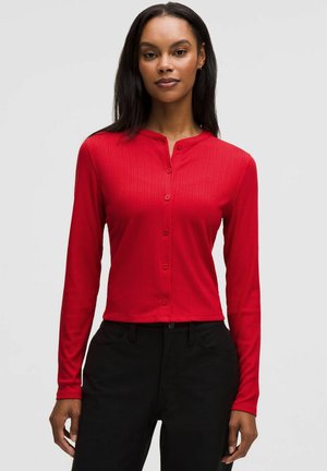 Vrouw met een aansluitend rood overhemd met knopen en lange mouwen en een zwarte broek met hoge taille, staand tegen een effen lichte achtergrond.