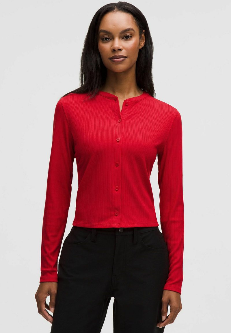 Femeie purtând un top roșu, cu mâneci lungi și nasturi, cambrat, și pantaloni negri cu talie înaltă, stând în picioare pe un fundal simplu și deschis la culoare.