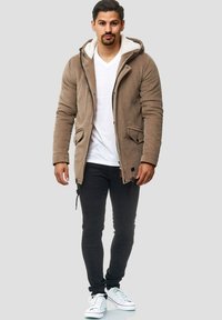Mand iført en tan kordfløjljakke med fleeceforing, hvid V-hals t-shirt, sorte tætsiddende bukser og hvide sneakers, står op mod en lys baggrund.