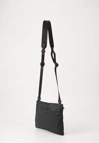 Svart crossover-veske laget av nylon med en glatt tekstur, som har en justerbar stropp og en glidelommer på baksiden.