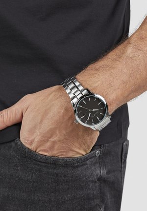 Silberne Armbanduhr aus Edelstahl mit schwarzem Zifferblatt, silbernen Stundenmarkierungen und einem Metallgliederarmband. Getragen an einem Handgelenk mit grauem Hemd und Jeans.