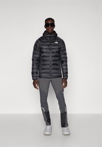 Schwarze Pufferjacke mit vertikalem Steppdesign und Kapuze, kombiniert mit grauen Sporthosen mit einem Muster und Akzenten. Weiße Sneakers.