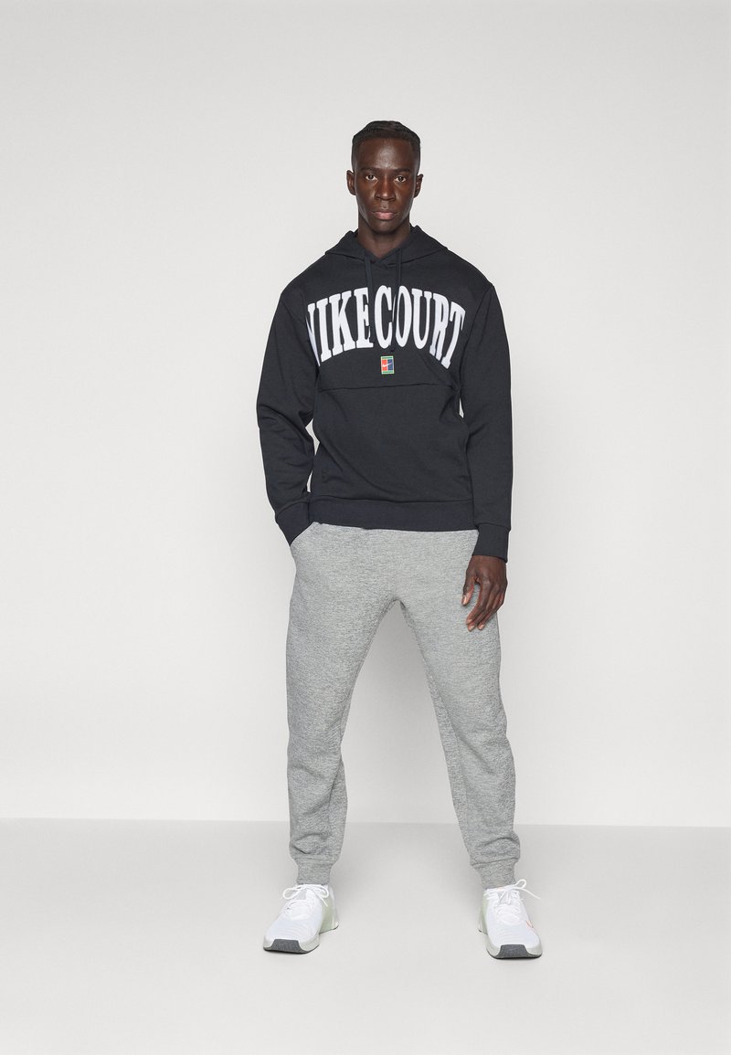 Felpa con cappuccio nera con la scritta "NIKECOURT" in bianco, abbinata a pantaloni della tuta grigi chiari e sneakers bianche con dettagli grigi.