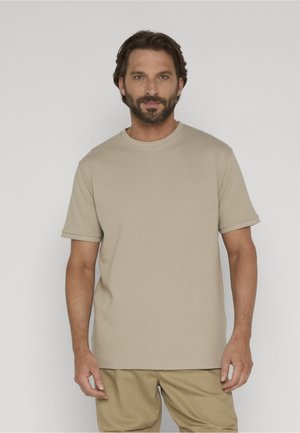 Uomo con capelli corti e scuri e barba, indossa una camicia beige a maniche corte e pantaloni coordinati, in piedi contro uno sfondo chiaro uniforme.