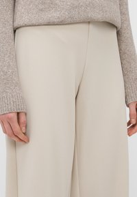 Pantaloni larghi beige realizzati in un tessuto liscio, con design pulito e texture sottile. Mano mostrata appoggiata sul tessuto.
