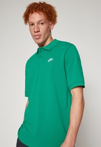 Vihreä puuvillapaita, jossa on klassinen kaulus, lyhyet hihat ja vasemmassa rinnassa valkoinen Nike-logot. Pehmeä rakenne.