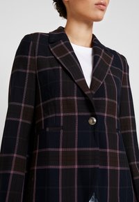 Granatowa marynarka w kratę z jasnoróżowymi i brązowymi wzorami, pojedynczy srebrny guzik oraz klapy z wycięciami. Gładka tkanina o dopasowanym kroju.