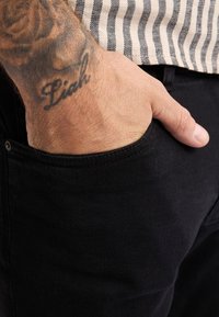 Jeans in denim nero con una tasca anteriore, mano appoggiata all'interno. Pelle visibile sul polso con un tatuaggio che recita "Liah". Tessuto a righe sopra.