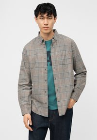 Νεαρός άνδρας που φοράει πουκάμισο με καρό κουμπιά πάνω από μια τιρκουάζ μπλουζά με γραφικά και σκούρο τζιν, όρθιος με το ένα χέρι στην τσέπη.