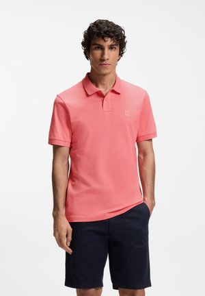 Férfi korallrózsaszín rövid ujjú galléros pólóingben és sötét tengerészkék rövidnadrágban, fehér háttér előtt áll, egyik kezét zsebre dugva.