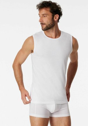 Mann mit lockigem Haar und Bart, der ein weißes ärmelloses Unterhemd und weiße Boxershorts trägt und nach links schaut.