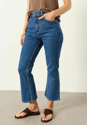 Femme portant un jean bleu coupé droit, un haut en knit marron et des sandales tressées marron, debout avec une main dans la poche.