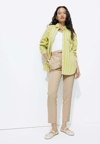 Chemise à rayures vertes et blanches superposée à un débardeur blanc, associée à un pantalon beige et des chaussures blanches avec des taches brunes.