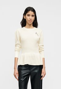 Εκρού πουλόβερ πλεκτό με ριμπ ύφανση, πεπλόμ, μακριά μανίκια, στρογγυλή λαιμόκοψη και διακοσμητικό καρφίτσα με λουλούδια· συνδυασμένο με μαύρο παντελόνι από ψεύτικο δέρμα.