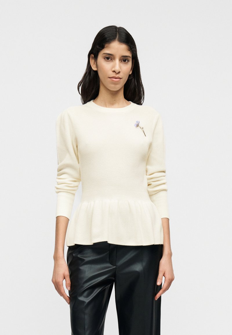 Εκρού πουλόβερ πλεκτό με ριμπ ύφανση, πεπλόμ, μακριά μανίκια, στρογγυλή λαιμόκοψη και διακοσμητικό καρφίτσα με λουλούδια· συνδυασμένο με μαύρο παντελόνι από ψεύτικο δέρμα.